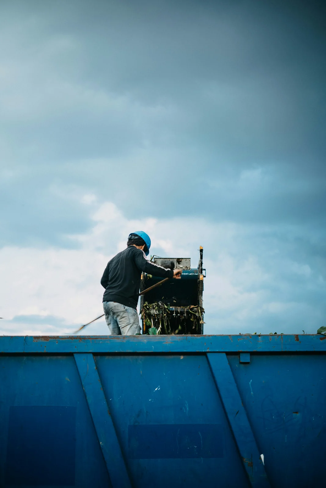 A worker levelling a load on top of a blue roll-off container